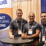 Three men in gray shirts smile at the camera in an expo.